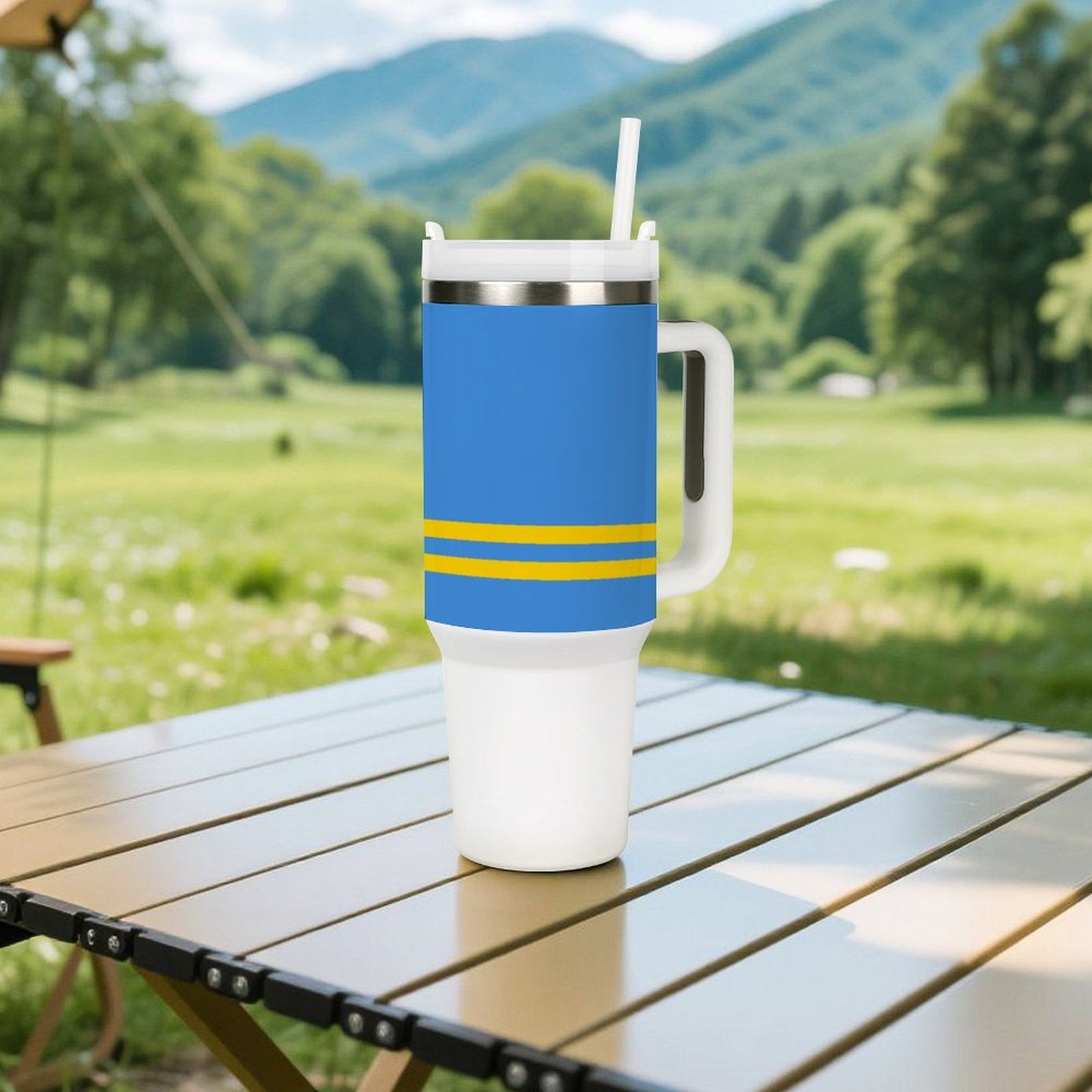 Blue [Brand] tumbler with handle on outdoor table, mountains backdrop.