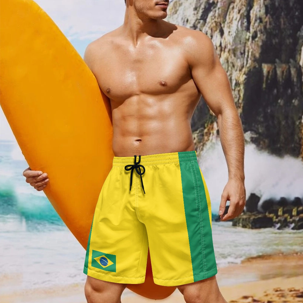 Brazil flag inspired beach shorts with yellow and green panels shown on model holding surfboard at tropical beach